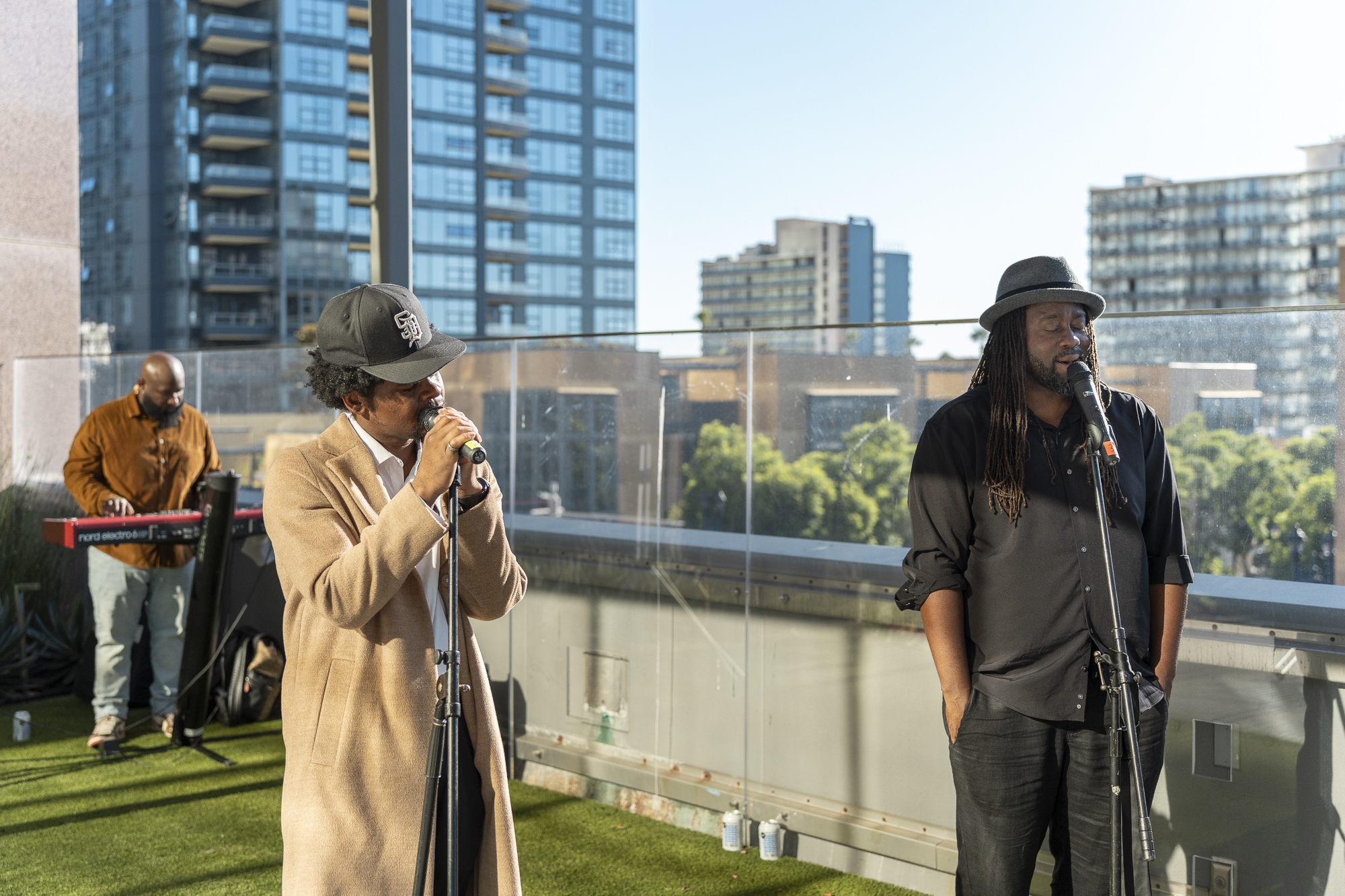 Two men with hats on sing in to microphones. A third man in the background plays a keyboard.