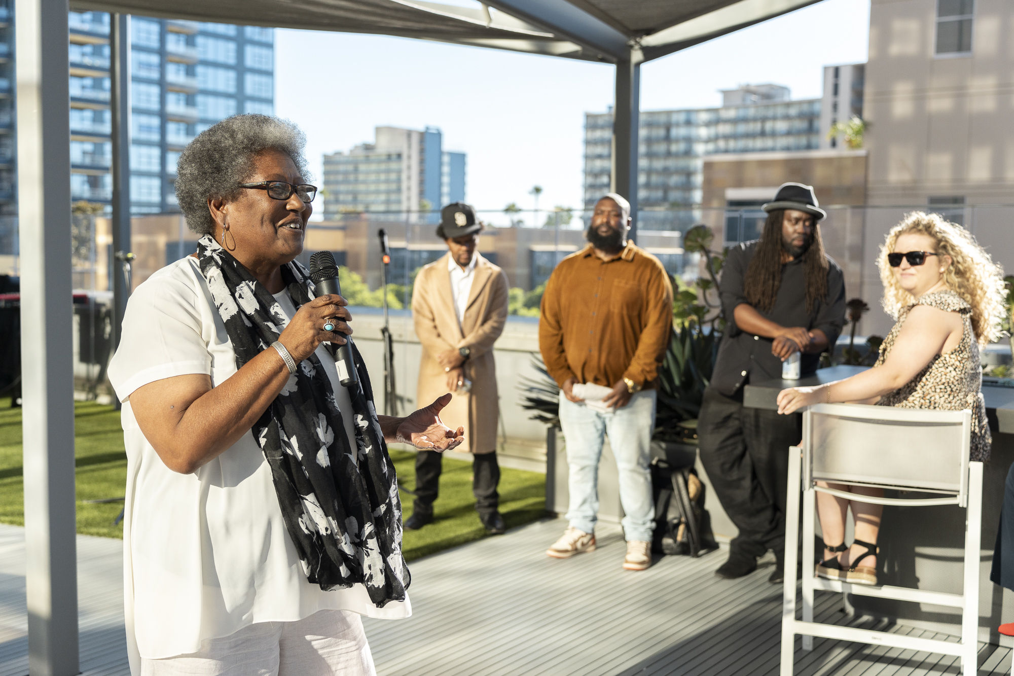 A woman holds a microphone and speaks to a crowd. 4 onlookers stand behind her listening.